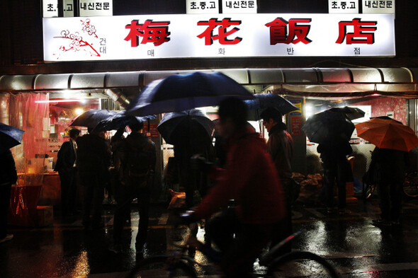 사람들이 비가 오는 날인데도 꼬치를 먹으려고 음식점 앞에 줄을 서서 기다리고 있다.