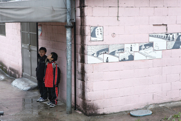 배다리 골목길의 한 처마 아래서 아이들이 빗줄기를 피하고 있다. 건물 벽면에 배다리 관통 산업도로 개설에 반대하는 예술가들이 그려놓은 벽화가 보인다.
