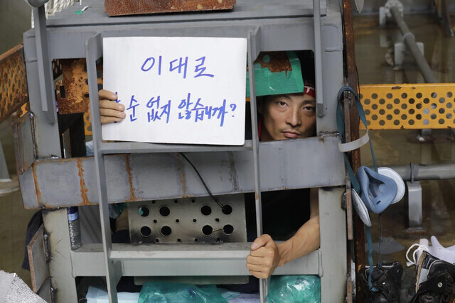 Yu Choe-an, the vice president of the shipbuilding subcontractor chapter of the KMWU, holds up a sign from his sit-in perch of a 1-square-meter metal box that has been welded shut. His sign reads, “Can’t you see this is unlivable?” (courtesy KMWU press center)