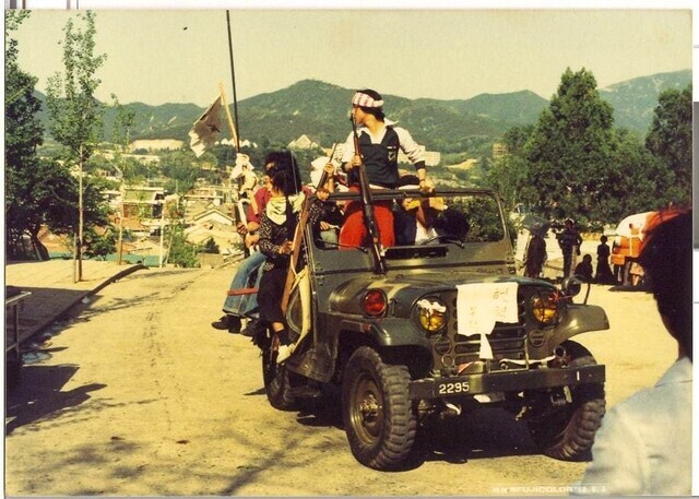 A vehicle driven by the Gwangju citizen militia sits atop a hill, with the city center visible in the background.