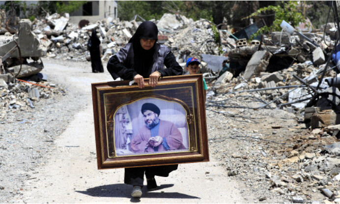 Uma mulher caminha entre casas destruídas segurando uma foto do líder do Hezbollah, Hassan Nasrallah, em 29 de junho (hora local), na vila de Aita al-Shaab, no sul do Líbano, que foi destruída por um ataque aéreo israelense. AP Yonhap Notícias