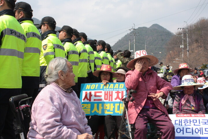 Elderly women sit on a road as trucks pass by