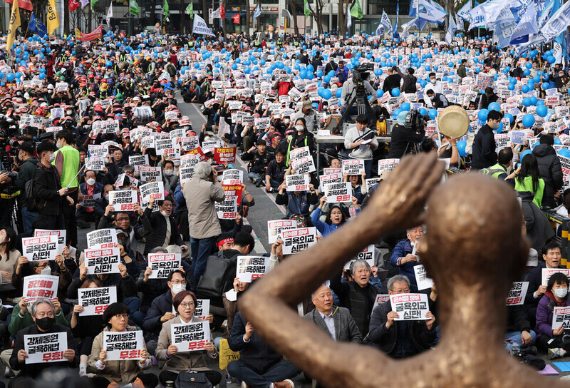 Participants in a nationwide rally on March 11 condemning Seoul’s “degrading” solution to forced mobilization compensation and calling for apology and compensation by Japan hold up signs as they sit in front of a statue in Seoul City Hall’s plaza representing victims of forced labor. (Kim Hye-yun/The Hankyoreh)