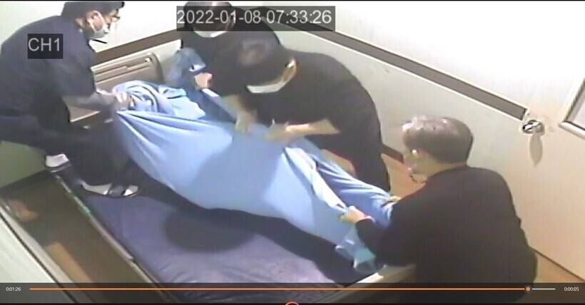 Still from CCTV footage showing orderlies wrapping up the deceased patient in a blanket for removal after he died while restrained in his bed. 