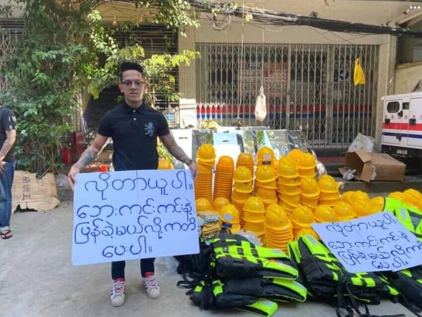 A Myanmar resident holding a sign that says, “take what you need and promise to come back alive,” offers plastic helmets and protective vests for free to anti-coup protesters. (Facebook screenshot)