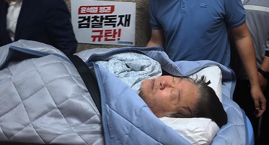 Democratic Party leader Lee Jae-myung is transported to a hospital on Sept. 18 due to deteriorating health amid a hunger strike. A sign next to his bed reads “We condemn the Yoon Suk-yeol regime’s prosecutor dictatorship!”