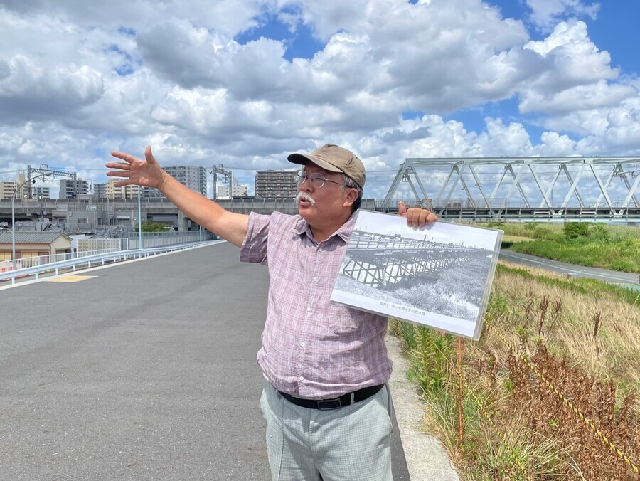 Masao Nishizaki, the director of the group Garden Balsam, stands at the Arakawa riverfront in Tokyo’s Sumida special ward holding up a photo in one hand as he speaks about the massacre of Koreans 100 years ago on Sept. 1, 1923. The bridge behind Nishizaki is the Keisei Oshiage Line. (Kim So-youn/The Hankyoreh)