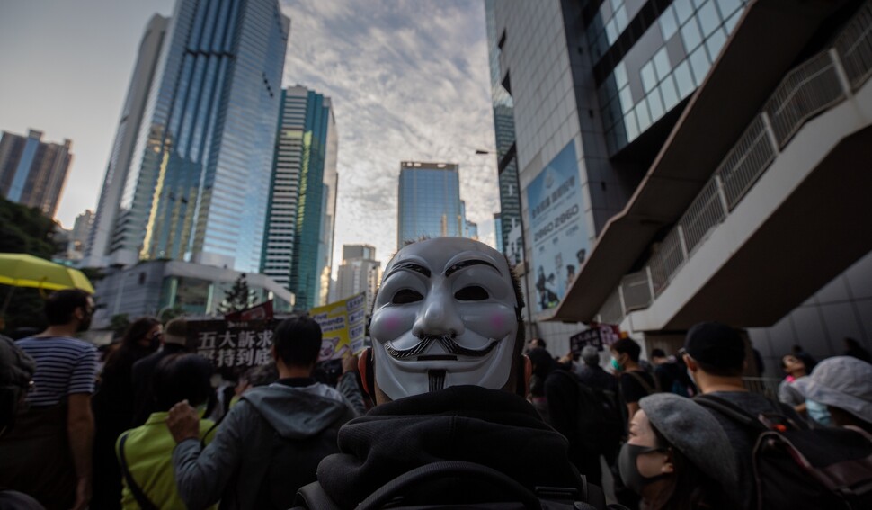 ‘가이 포크스’ 가면을 머리 뒤로 돌려쓴 시민이 시위대와 함께 행진하고 있다. 영화 <브이 포 벤데타>에 나온 이 가면은 저항의 상징으로 여겨진다.