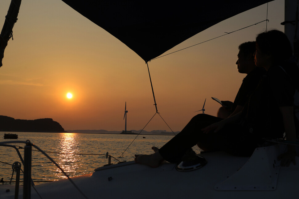 한 쌍의 젊은이가 뱃머리에 앉아 바다 저편으로 넘어가는 해를 바라보고 있다.