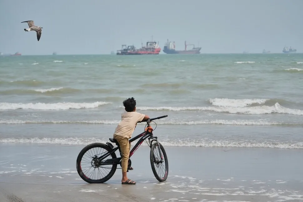 A boy stands with his bicycle on a beach overlooking commercial vessels anchored in the Strait of Hormuz off Bandar Abbas, Iran, Sept. 7, 2026. (AP/Yonhap)