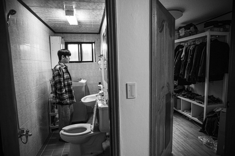 헤이든이 집에서 화장실 거울에 비친 자신의 옷차림을 보고 있다. 옆방에는 남성복이 잔뜩 걸려 있다.