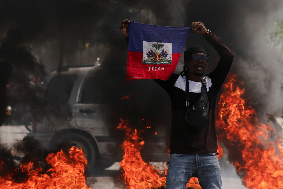2024년 3월1일 아이티 수도 포르토프랭스에서 아리엘 앙리 총리의 퇴진을 요구하는 시위가 벌어졌다. 화염에 휩싸인 거리에서 한 참가자가 아이티 국기를 들고 구호를 외치고 있다. REUTERS
