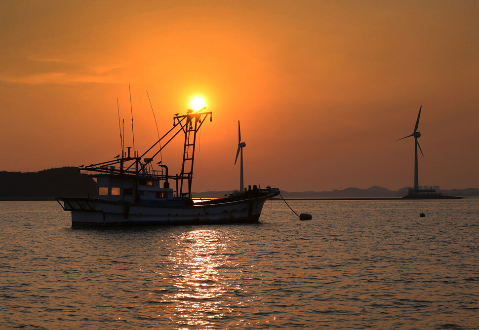 경기도 화성시 서신면 전곡항 앞바다에서 2025년 9월11일 누에섬과 풍력발전기 사이로 해가 넘어가는 동안 안강망 어선 한 척이 그 앞을 지나고 있다.