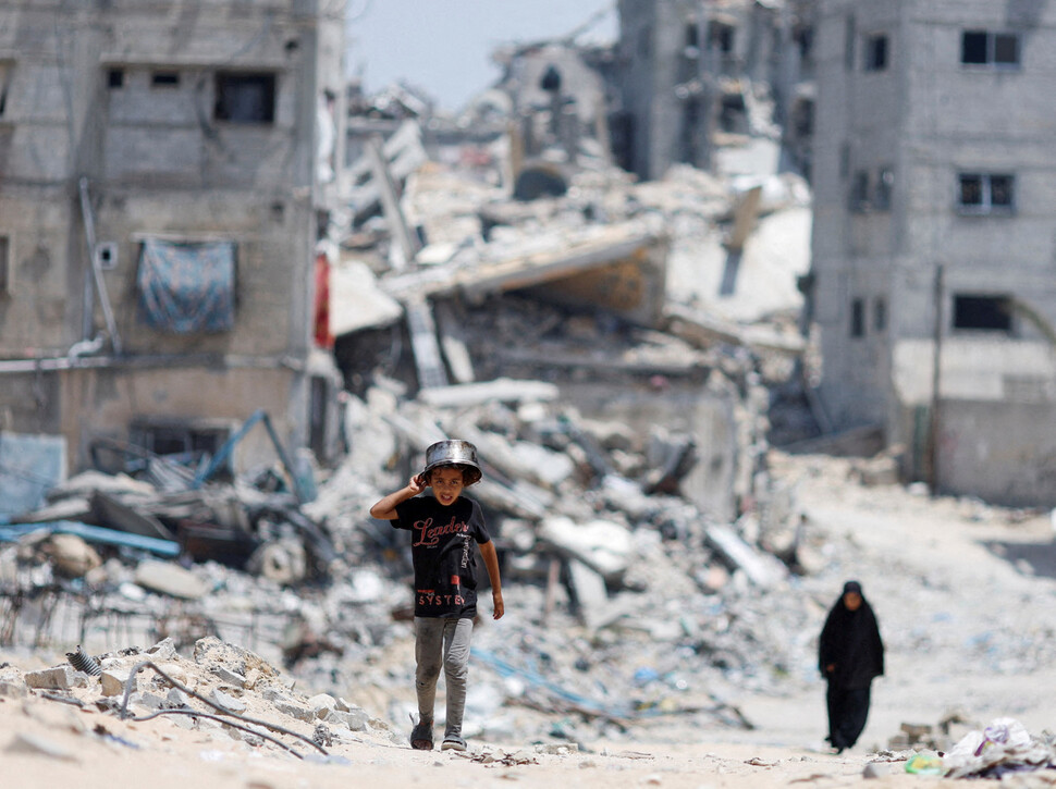 2024년 5월22일 팔레스타인 땅 가자지구 남쪽 칸유니스에서 한 어린이가 식량 배급을 받기 위해 솥단지를 머리에 이고 무너진 건물 더미가 즐비한 거리를 지나고 있다. REUTERS 연합뉴스