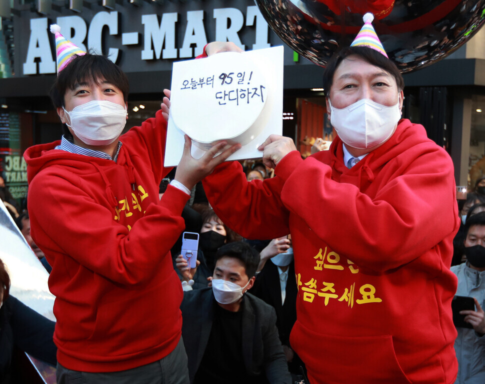 People Power Party leader Lee Jun-seok and the party’s presidential nominee Yoon Seok-youl pose for a photo holding up a cake for Yoon’s birthday on Dec. 4 at Seomyeon Young Street in Busan. The cake reads, “95 days left! Let’s do a great job.” (Yonhap News)